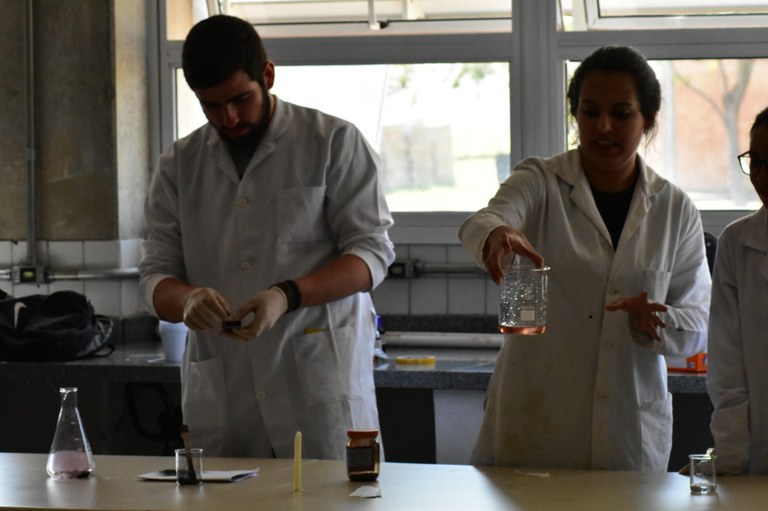 Três alunos de jaleco branco demonstram em cima de uma mesa o preparo de misturas químicas para realizar a demonstração de experimento forense para reconhecimento de digital.