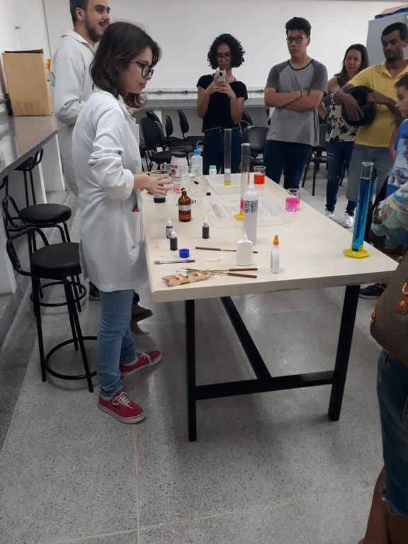 equipe de alunos de jaleco branco demonstram numa mesa um experimento lúdico chamado "pasta de elefante", onde a mistura de determinados produtos resulta numa reação química que forma uma espuma colorida dentro de um tubo de ensaio e esta vai subindo rápido até transbordar.