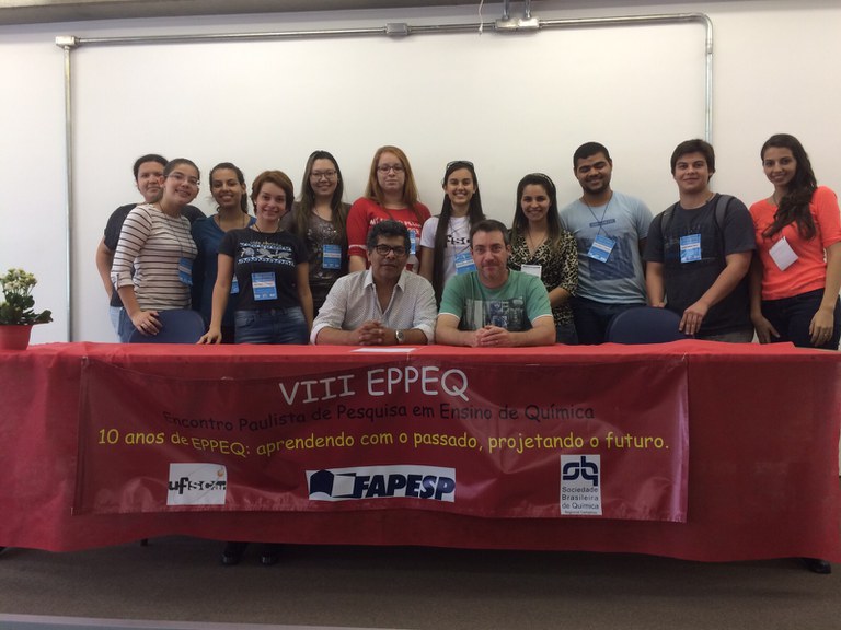 mesa retangular grande com uma toalha vermelha e uma faixa com o nome do evento VIII EPPEQ 2015, estão sendo à mesa Prof. Dr. João Batista dos Santos Junior e Prof. Dr. Edemar Benedetti Filho, atrás dos professores estão onze alunos do Curso de Graduação Licenciatura em Química, que fazem parte da comissão organizadora do evento.